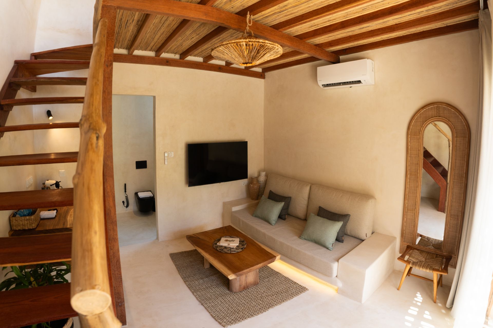 Interior of a small living space with a sofa, stairs, and a mirror.  Neutral colors and natural wood accents.