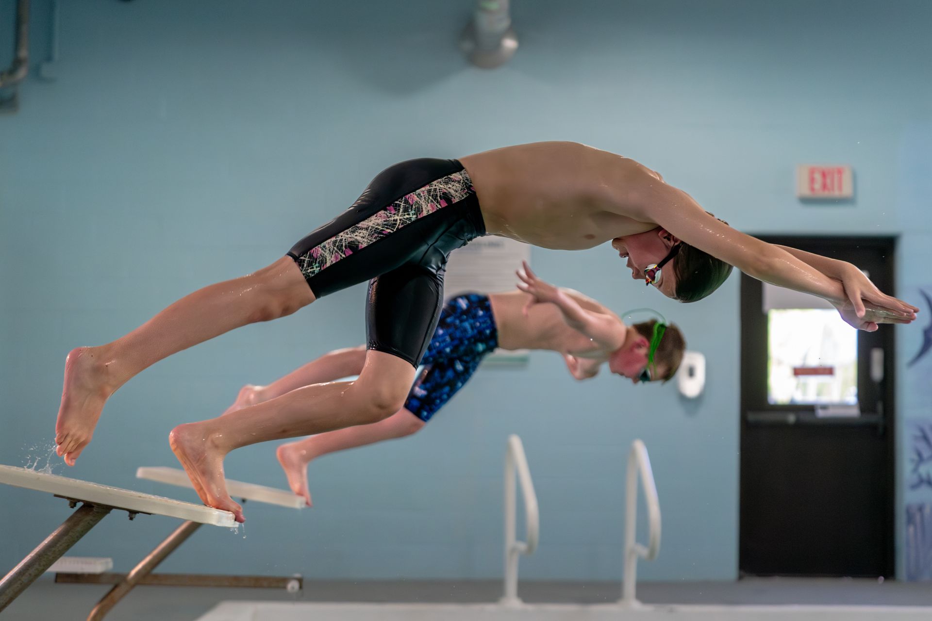 Two swimmers dive off starting blocks into a pool, midair in a blue indoor natatorium.