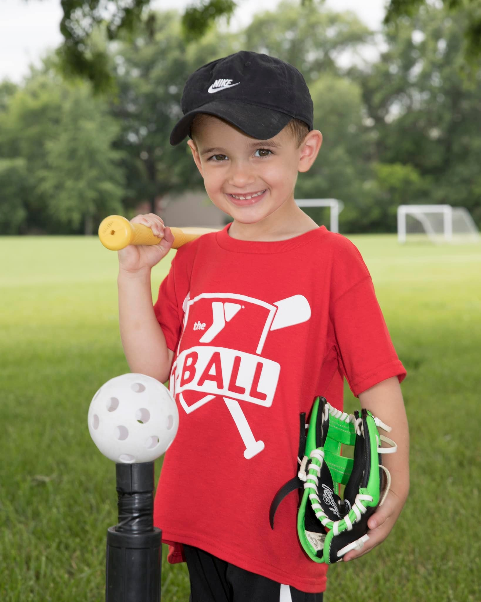 Young boy in baseball gear, smiling, holding bat, glove, and tee ball. Wearing red shirt and black hat, outside on a field.