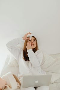 Woman applying eye drops, sitting on a bed with a laptop, wearing white sweater.
