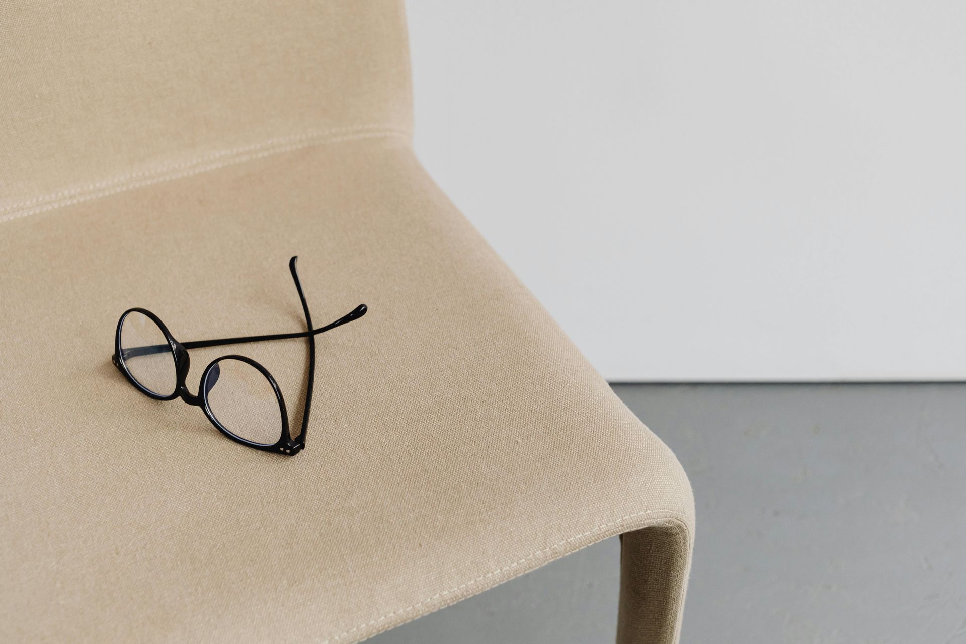 Pair of black wireframe eyeglasses resting on a beige chair. White wall in background.