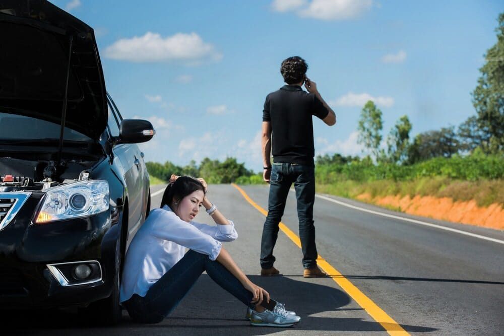 A Man And A Woman Are Sitting Next To A Broken Down Car On The Side Of The Road — Wilson's Towing In Withcott, QLD