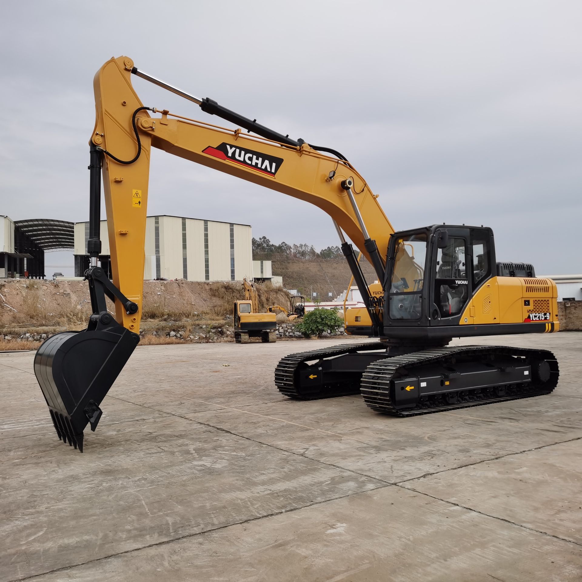 Yellow YUCHAI YC215 excavator on a concrete surface, boom extended. Construction site background.