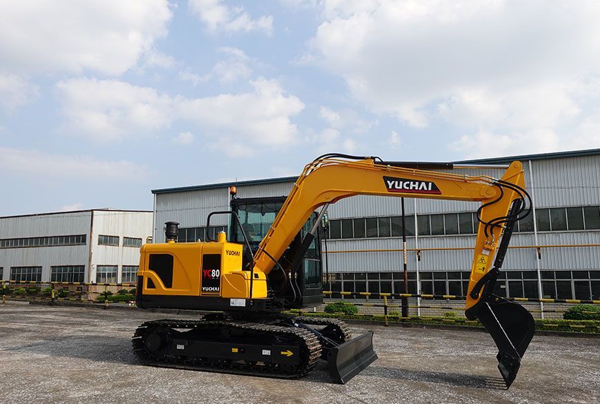 Yellow YUCHAI YC80 excavator on a concrete surface, parked near industrial buildings, under a cloudy sky.