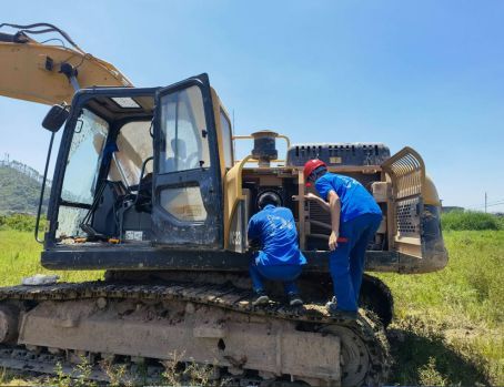 Two workers in blue uniforms repair a yellow YUCHAI excavator on muddy ground under a blue sky.