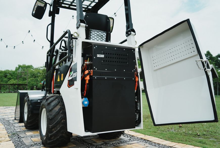 White and black compact loader with open side panel revealing battery and components, outdoors on grass.