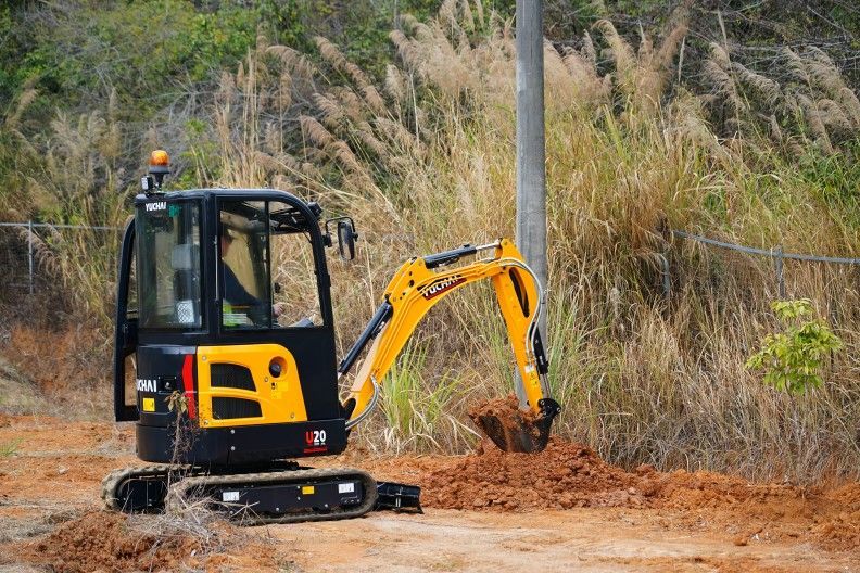 Une mini-pelle jaune et noire creuse la terre près d'une clôture.