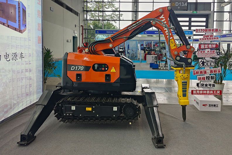 Robot de démolition orange et noir avec marteau-piqueur, en intérieur, jambes déployées pour plus de stabilité.