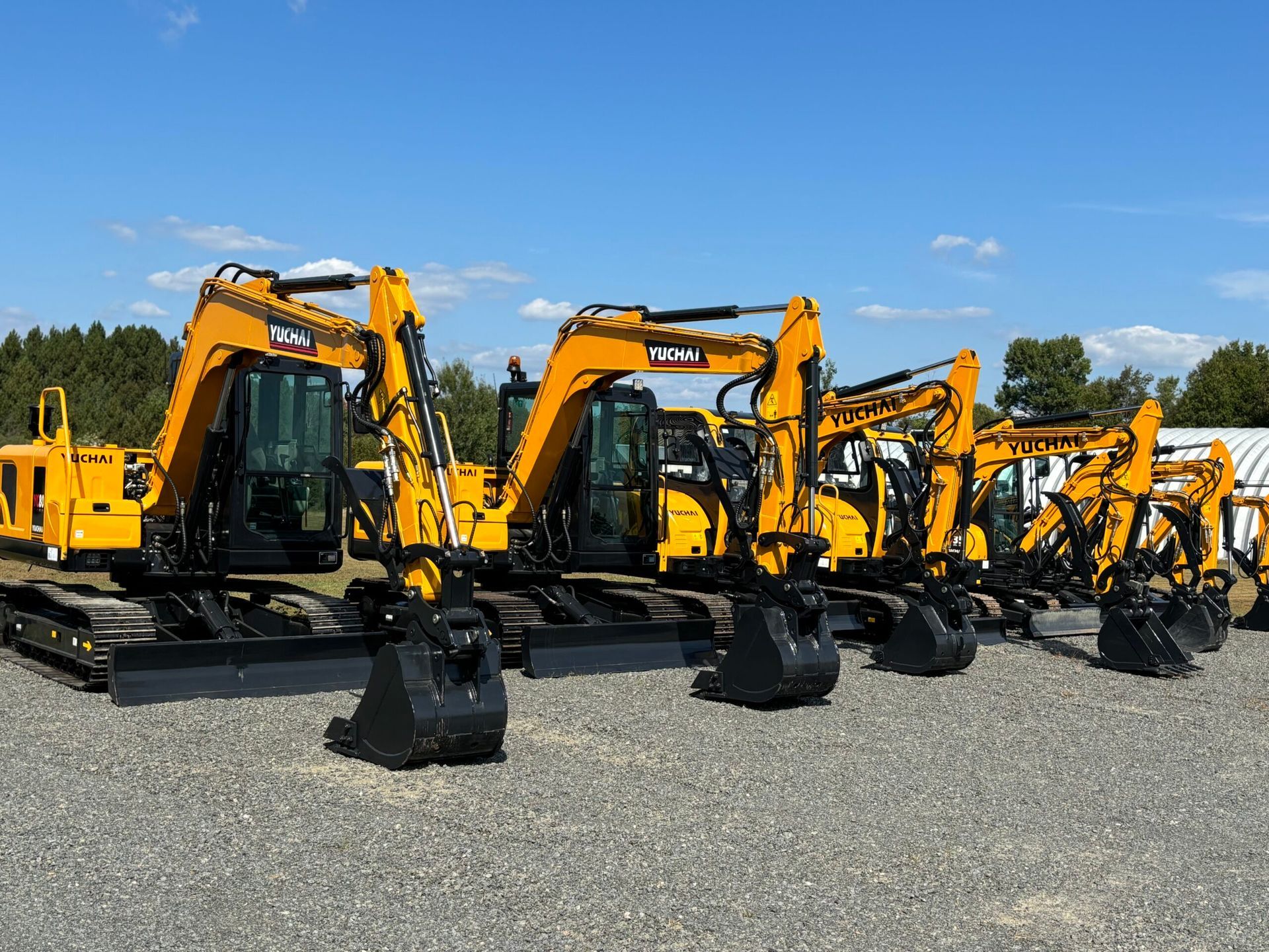 Several yellow YUCHAI excavators parked outdoors under a blue sky, on a gravel surface.