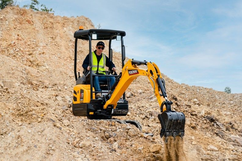 Un homme manœuvre une petite pelleteuse jaune sur un talus de terre.
