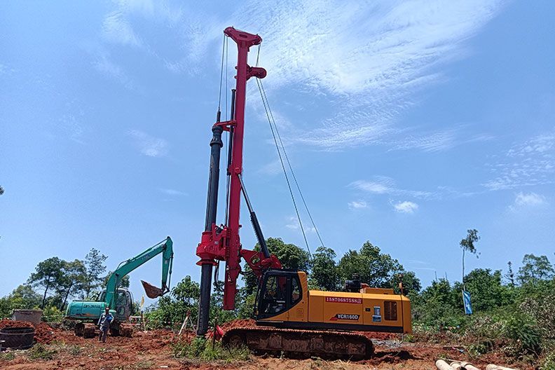 Construction site with a YUCHAI red drilling machine and green excavator under a blue sky.
