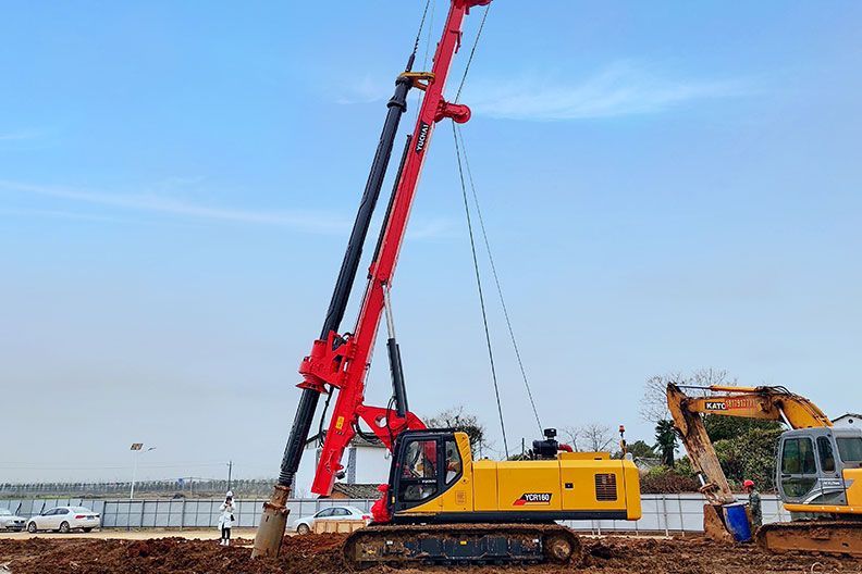 Red and yellow YUCHAI construction rig drilling into the ground, other machinery in the background, blue sky.