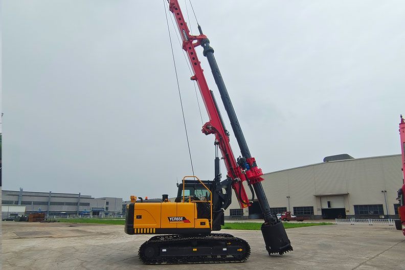 Red and yellow YUCHAI YCR65E drilling rig on tracks, angled against a cloudy sky, near a building.