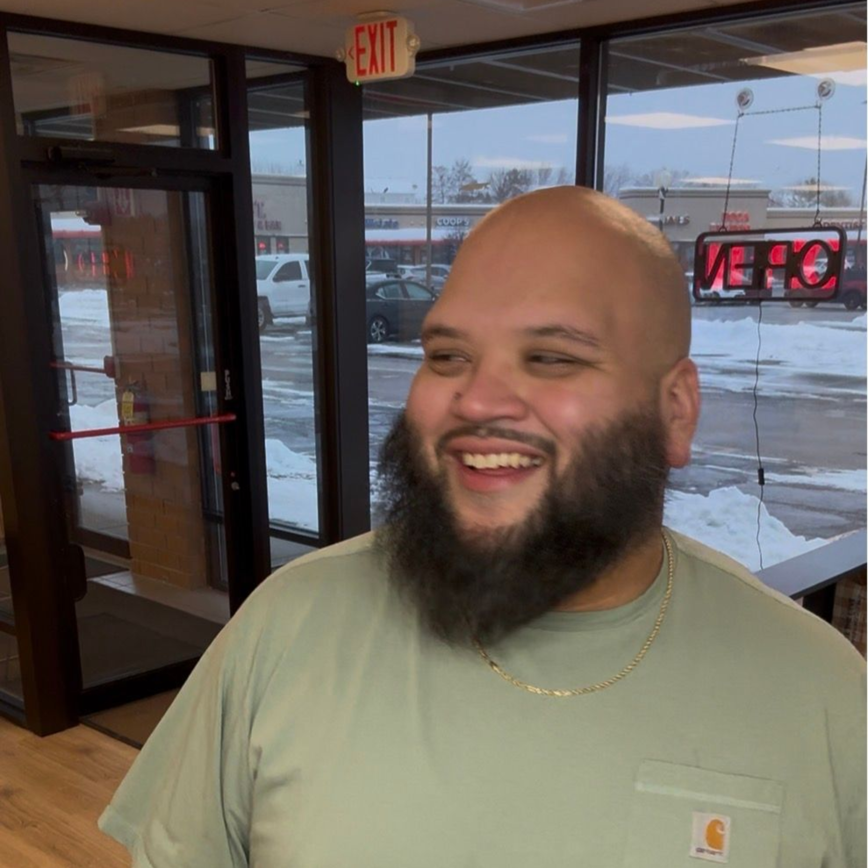 A smiling person with a beard and a shaved head, wearing a light green shirt, standing indoors near glass doors.