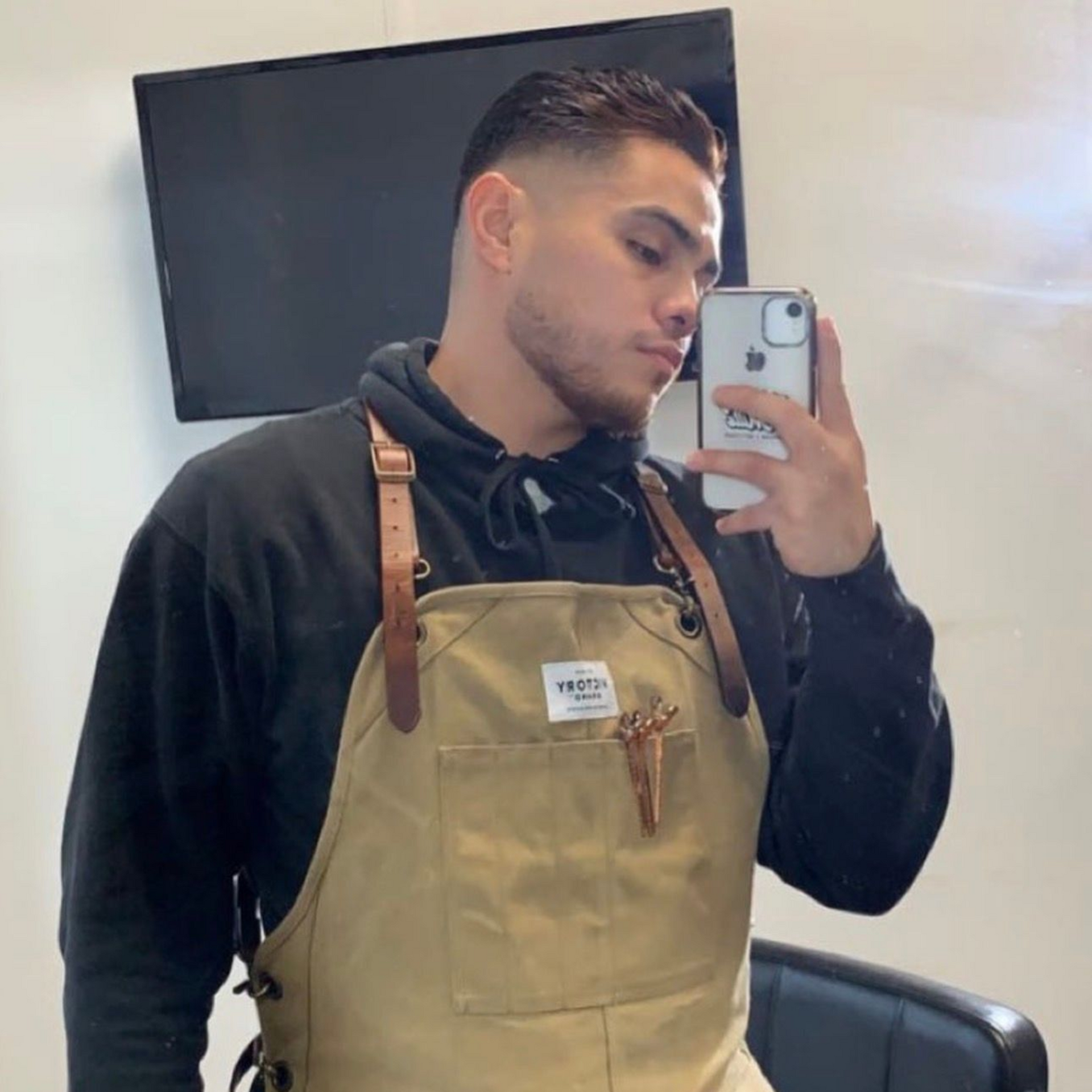 A barber in a tan apron and black hoodie takes a mirror selfie in a salon, showing his short, faded hairstyle.