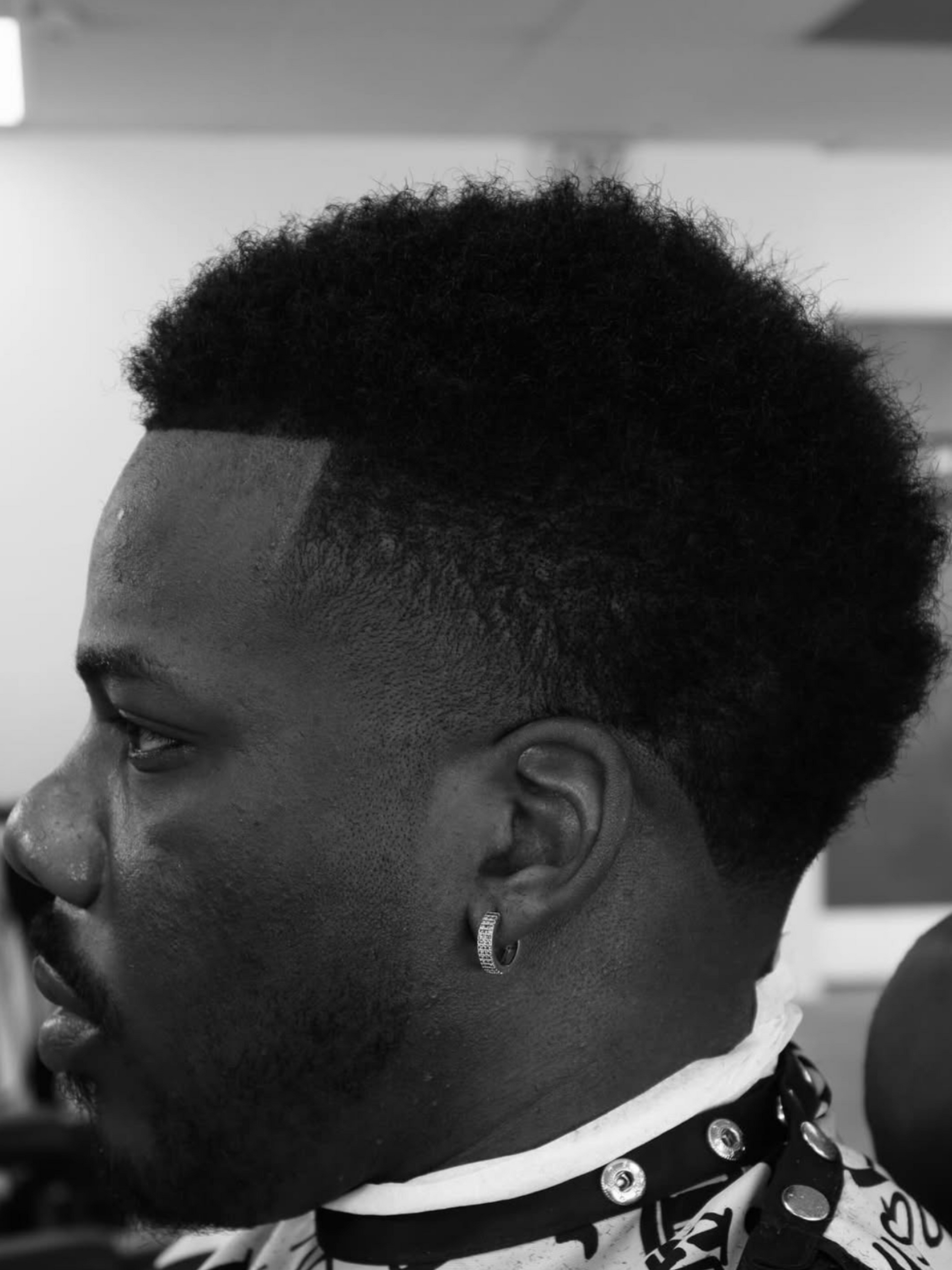 A side profile of a person with a short, textured, faded haircut and a sharp hairline, wearing a barber’s cape.