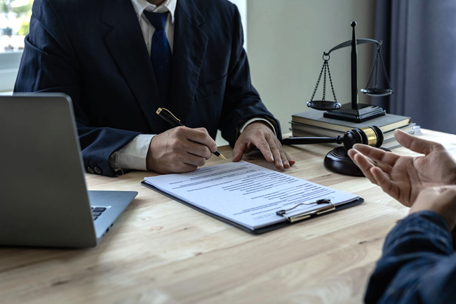Riunione di lavoro a una scrivania con un computer portatile, documenti legali, una bilancia della giustizia e due persone che discutono di un contratto.