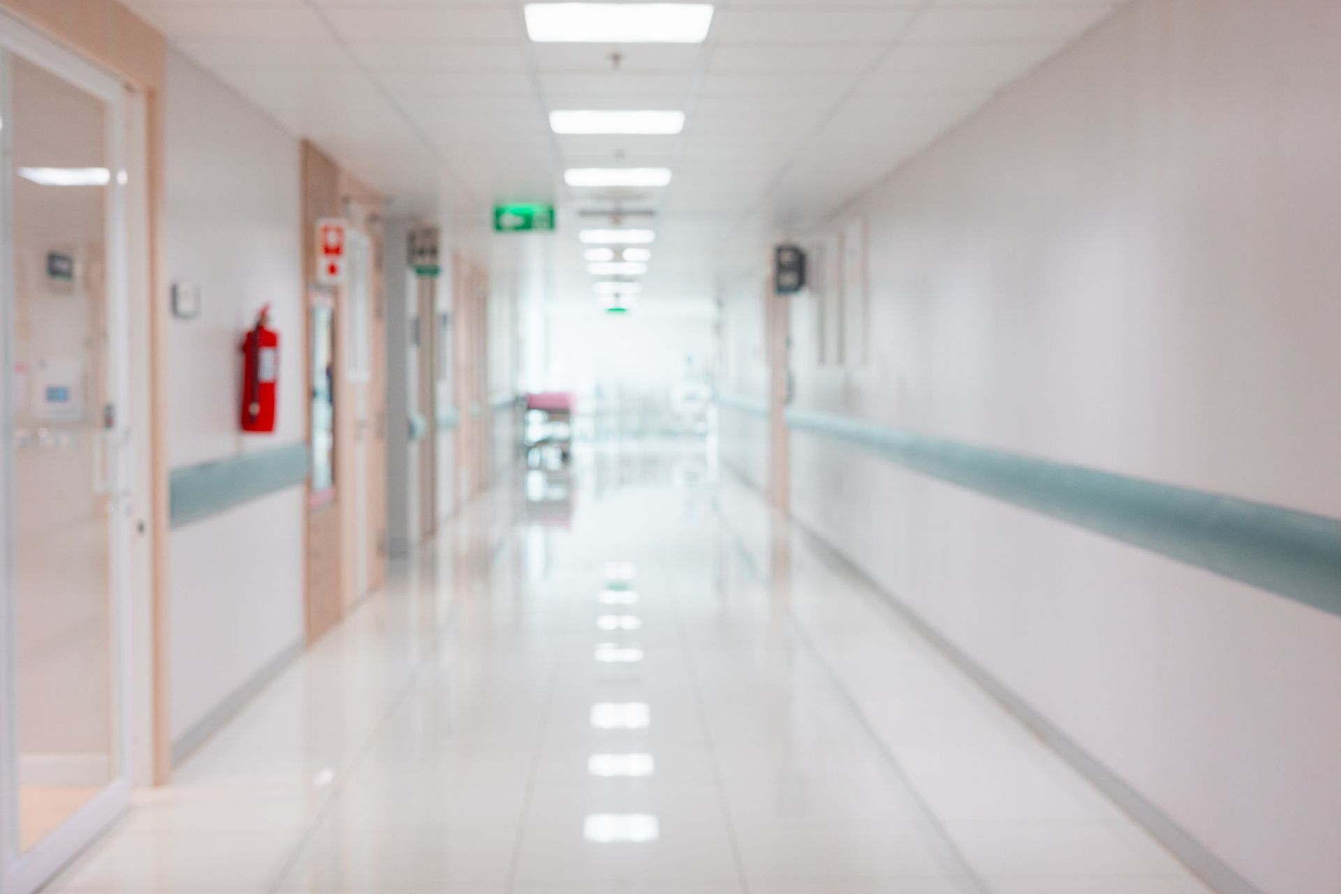 Blurred perspective of a hospital hallway with bright white walls, a fire extinguisher, and emergency exit signs. Blurred perspective of a hospital hallway with bright white walls, a fire extinguisher, and emergency exit signs.