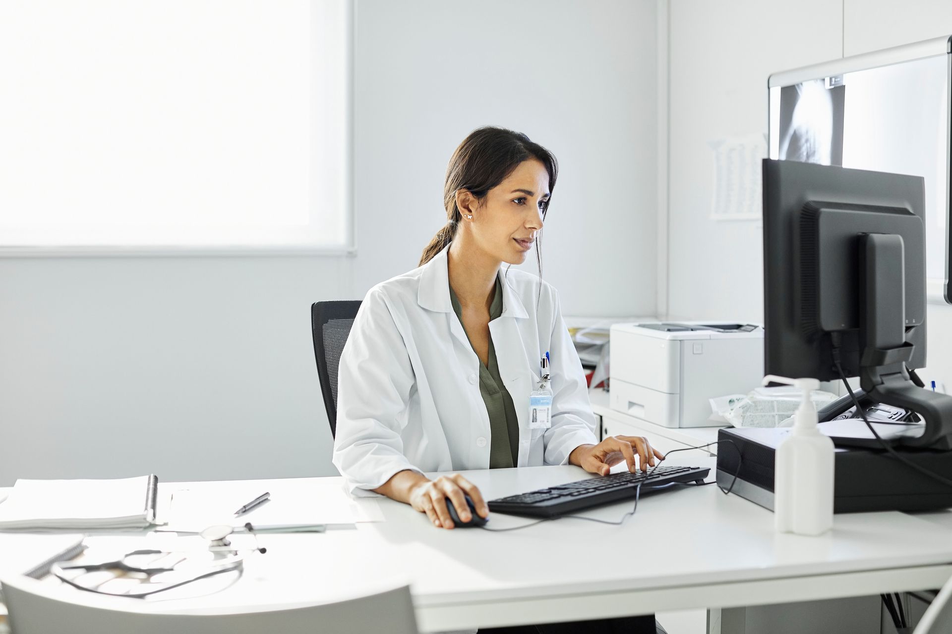 Doctor in a white coat working at a computer in a bright office.