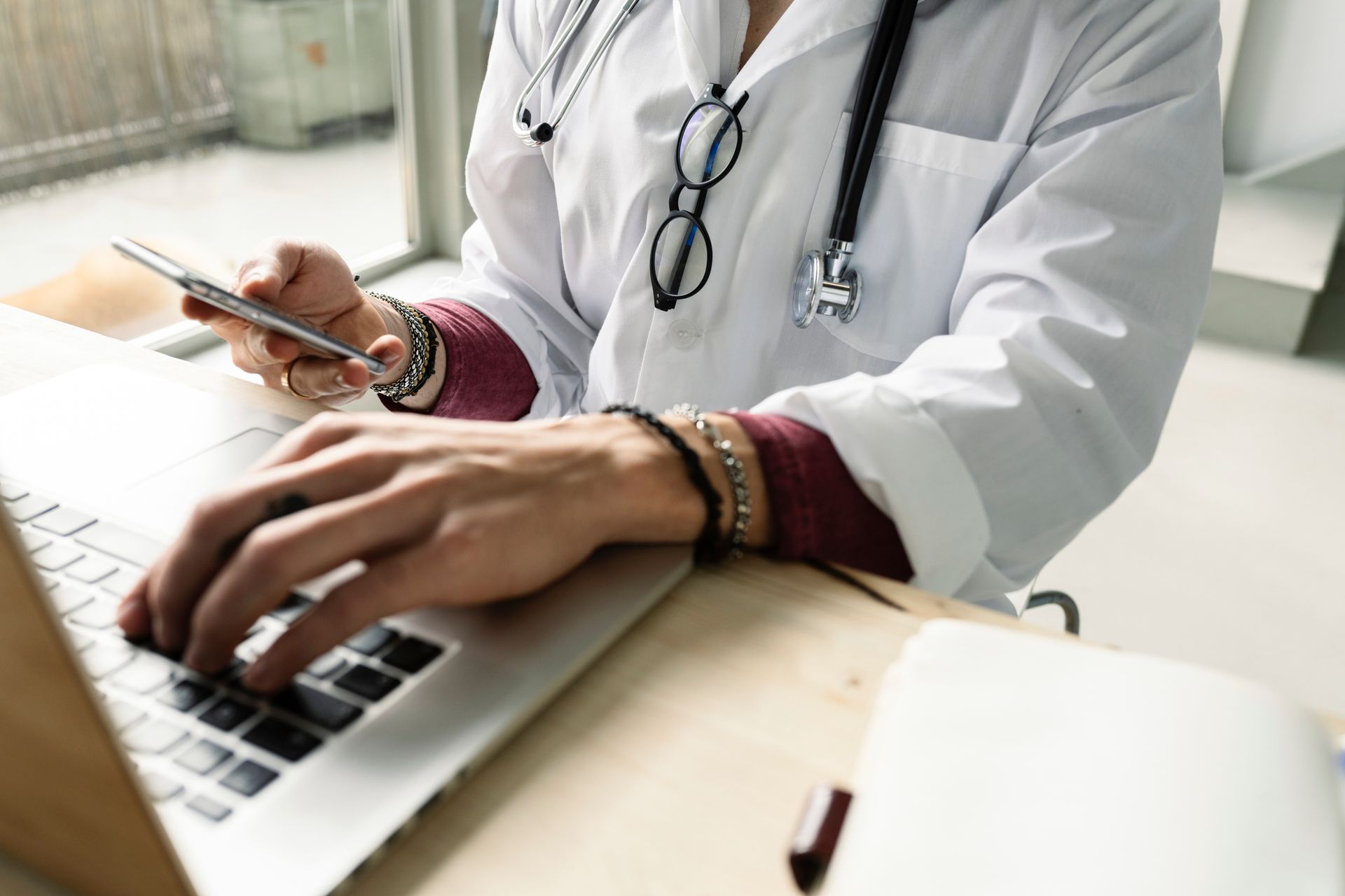 Doctor in white coat using laptop and cell phone, stethoscope around neck, glasses hanging. Doctor in white coat using laptop and cell phone, stethoscope around neck, glasses hanging.