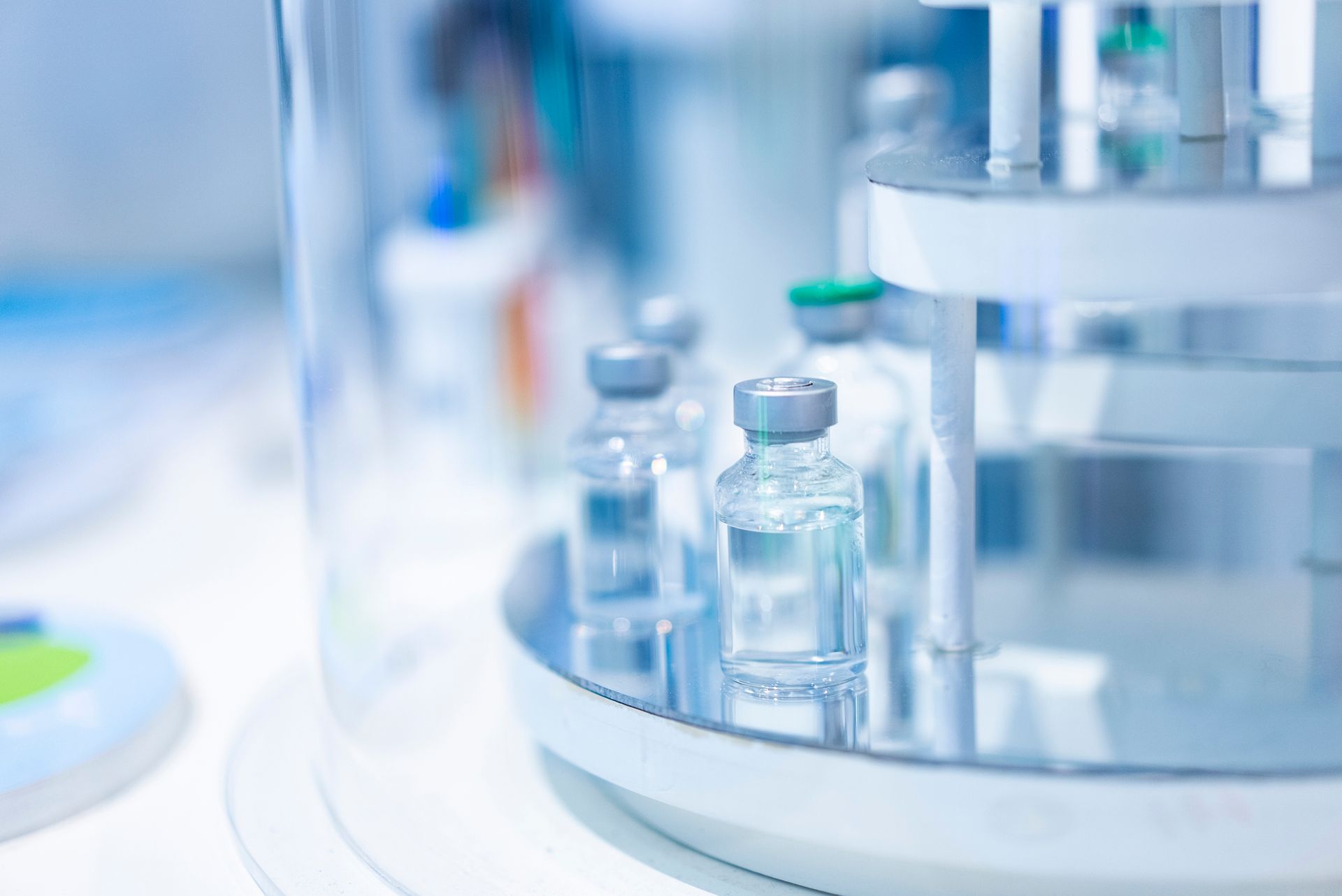 Glass vials with silver caps sit on a tray inside a laboratory machine. Glass vials with silver caps sit on a tray inside a laboratory machine.
