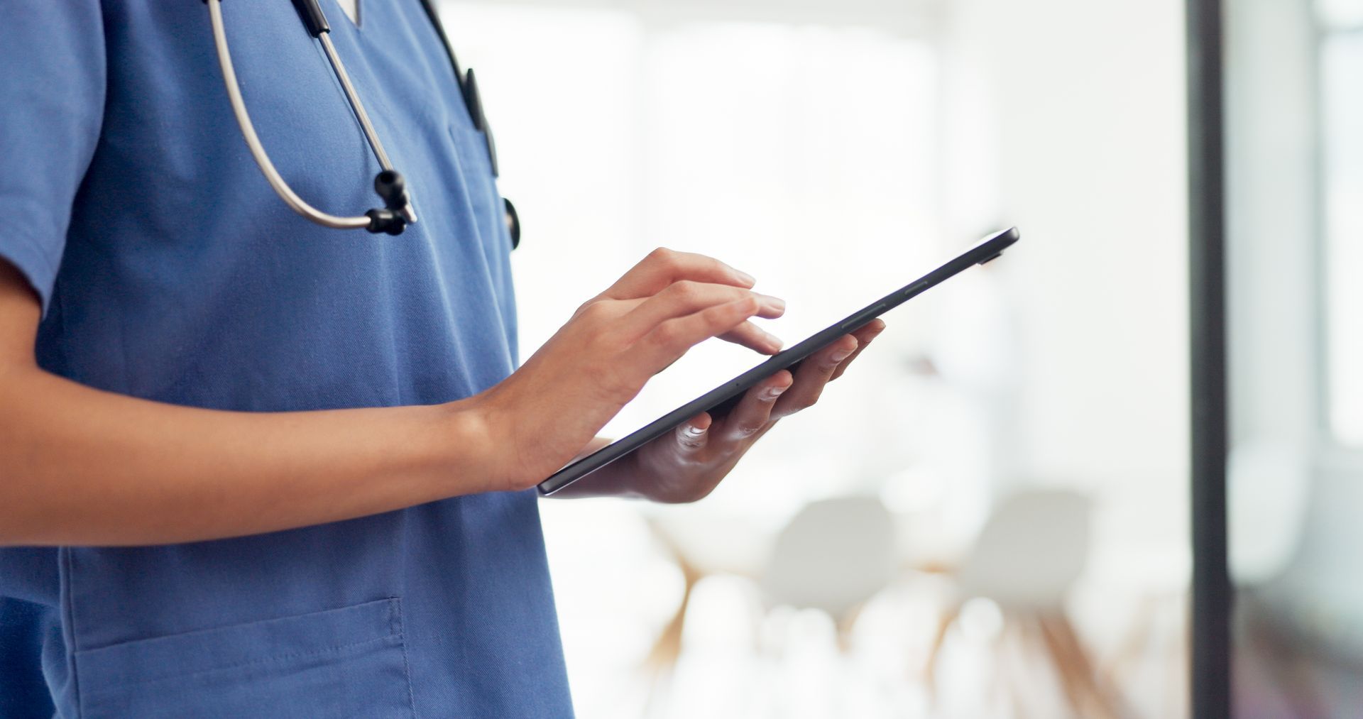 Healthcare worker in blue scrubs using a tablet, stethoscope around neck. Healthcare worker in blue scrubs using a tablet, stethoscope around neck.