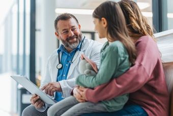 Doctor consulting with a child and parent in an office; doctor holds a tablet, smiling.
