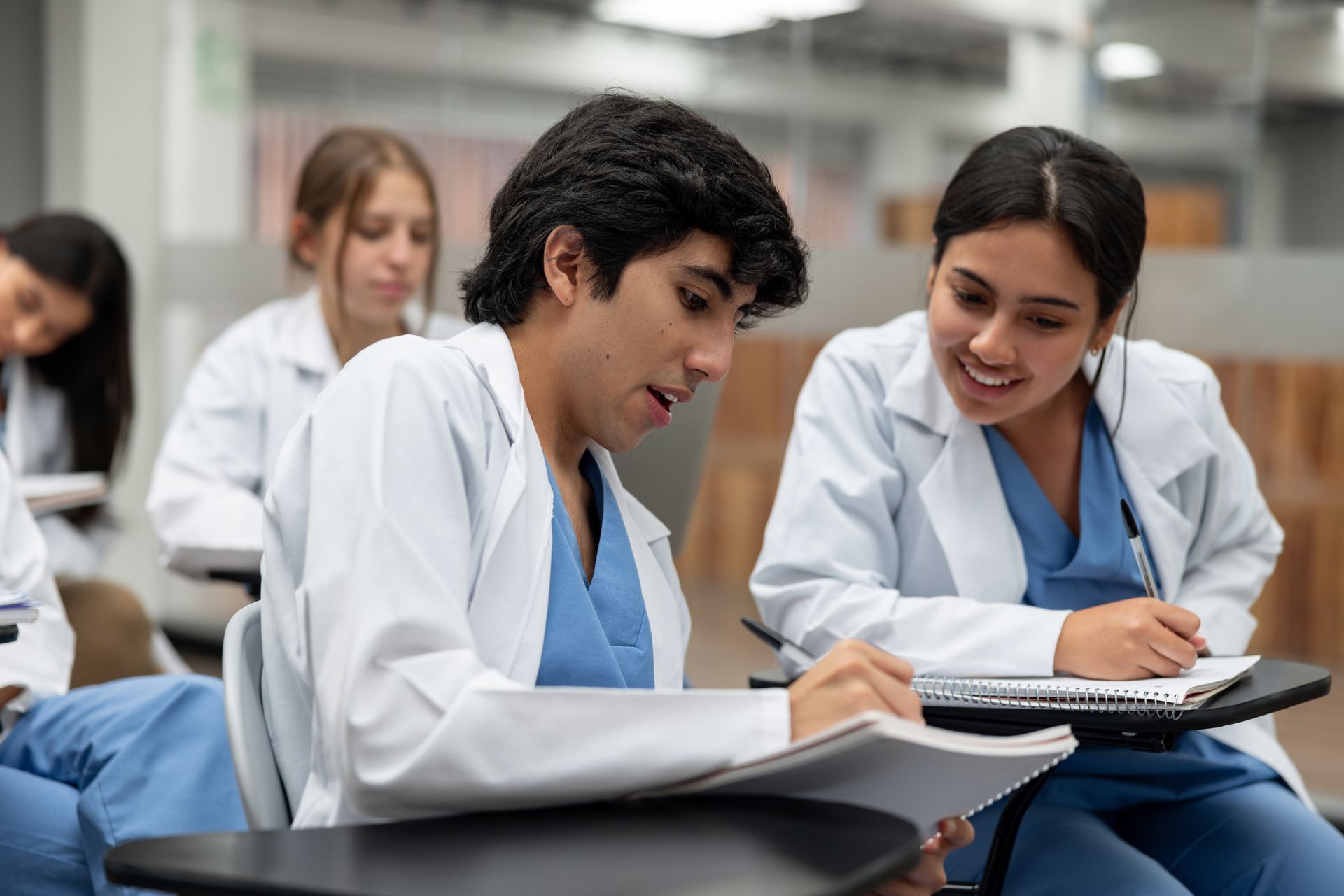 Two medical students in white coats study together in a classroom.