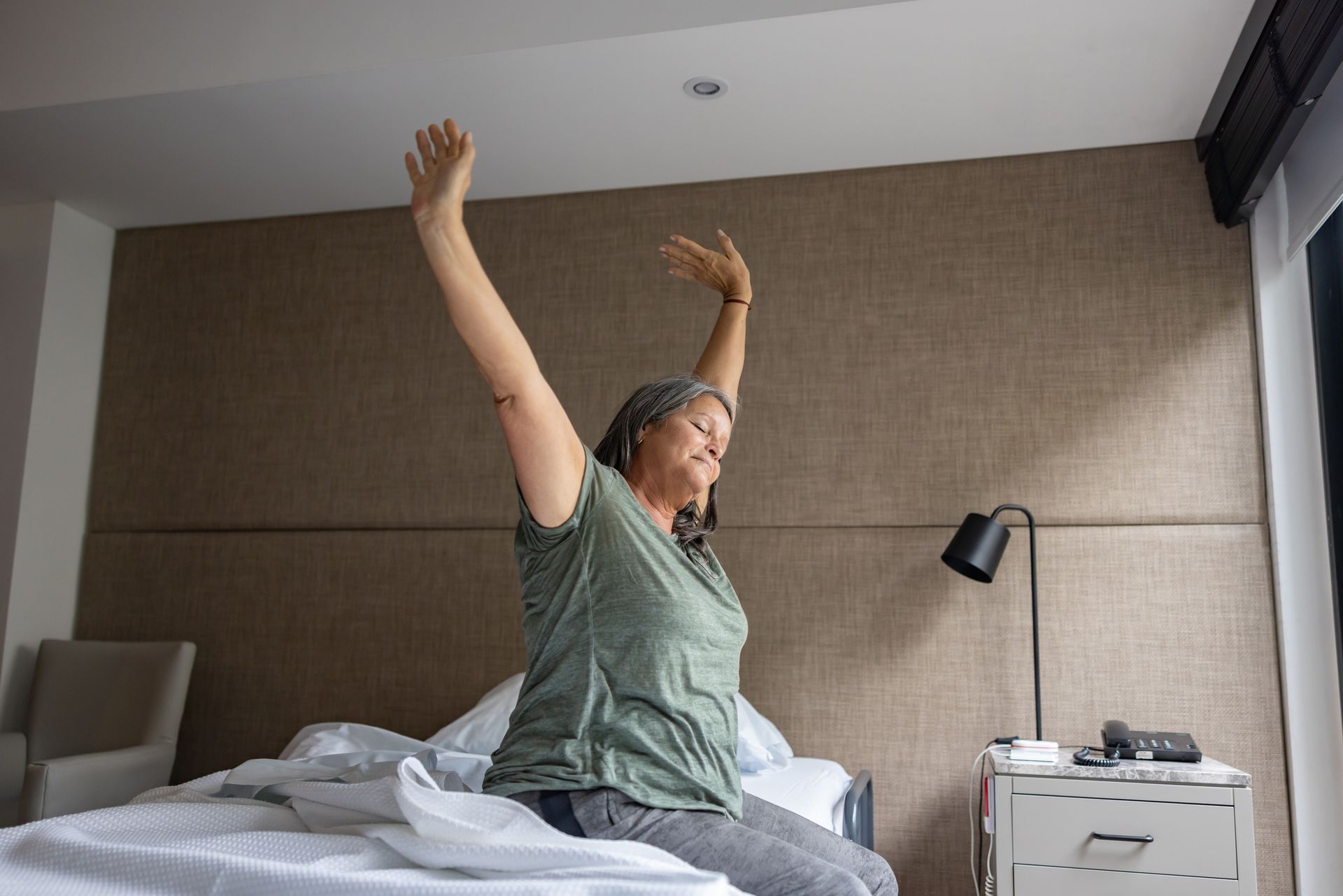 Woman sitting up in bed, stretching arms upward. Beige headboard in background.