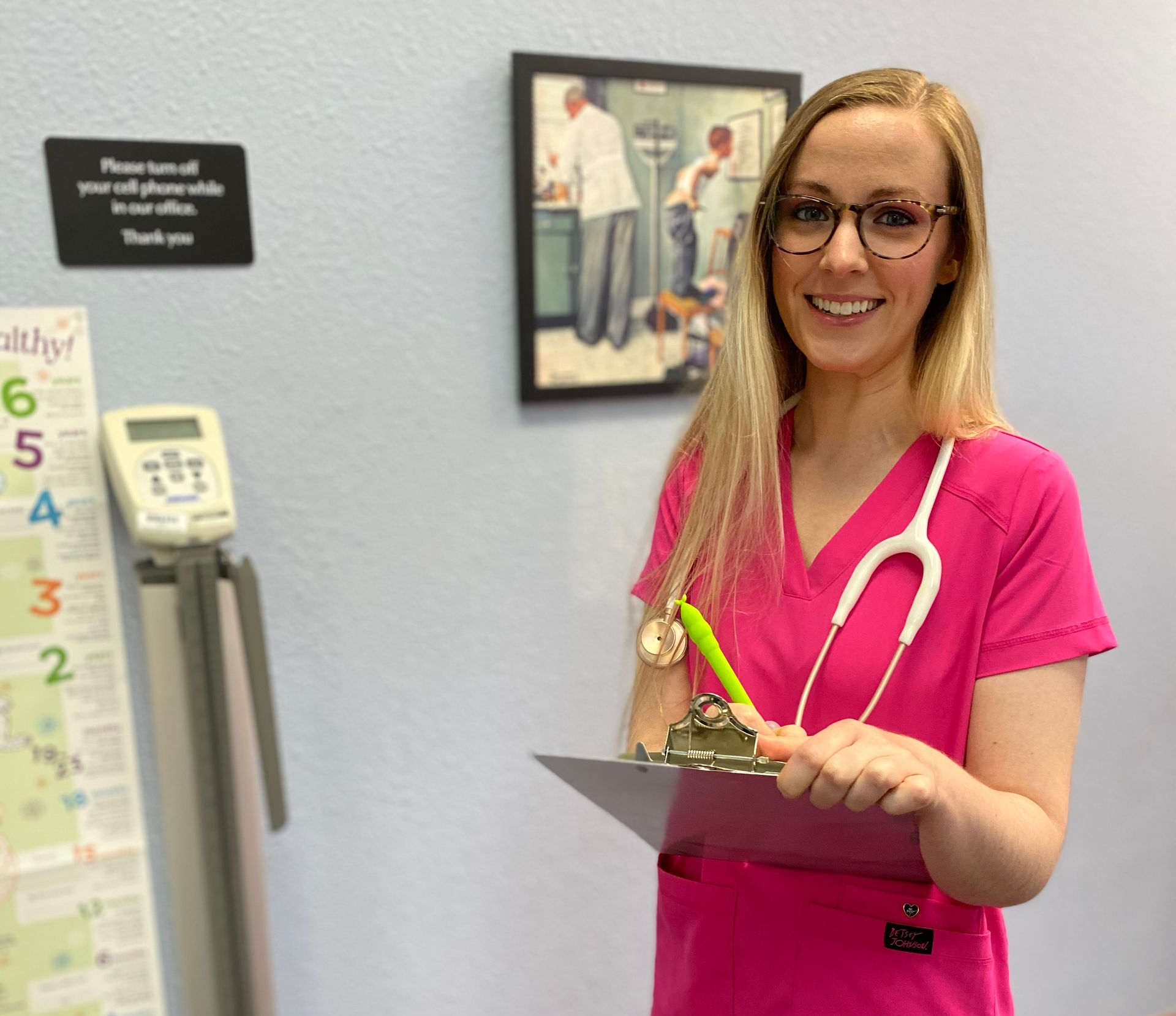 Healthcare worker in pink scrubs, holding a clipboard and stethoscope, smiling in a medical office setting.