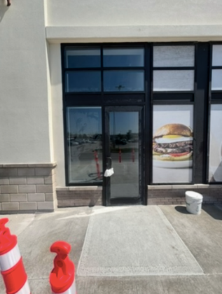 Storefront entrance with large windows and burger decal, flanked by traffic cones and a bucket on the sidewalk.