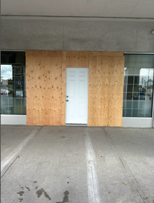 Boarded storefront entrance with a white door and windows under a concrete overhang.