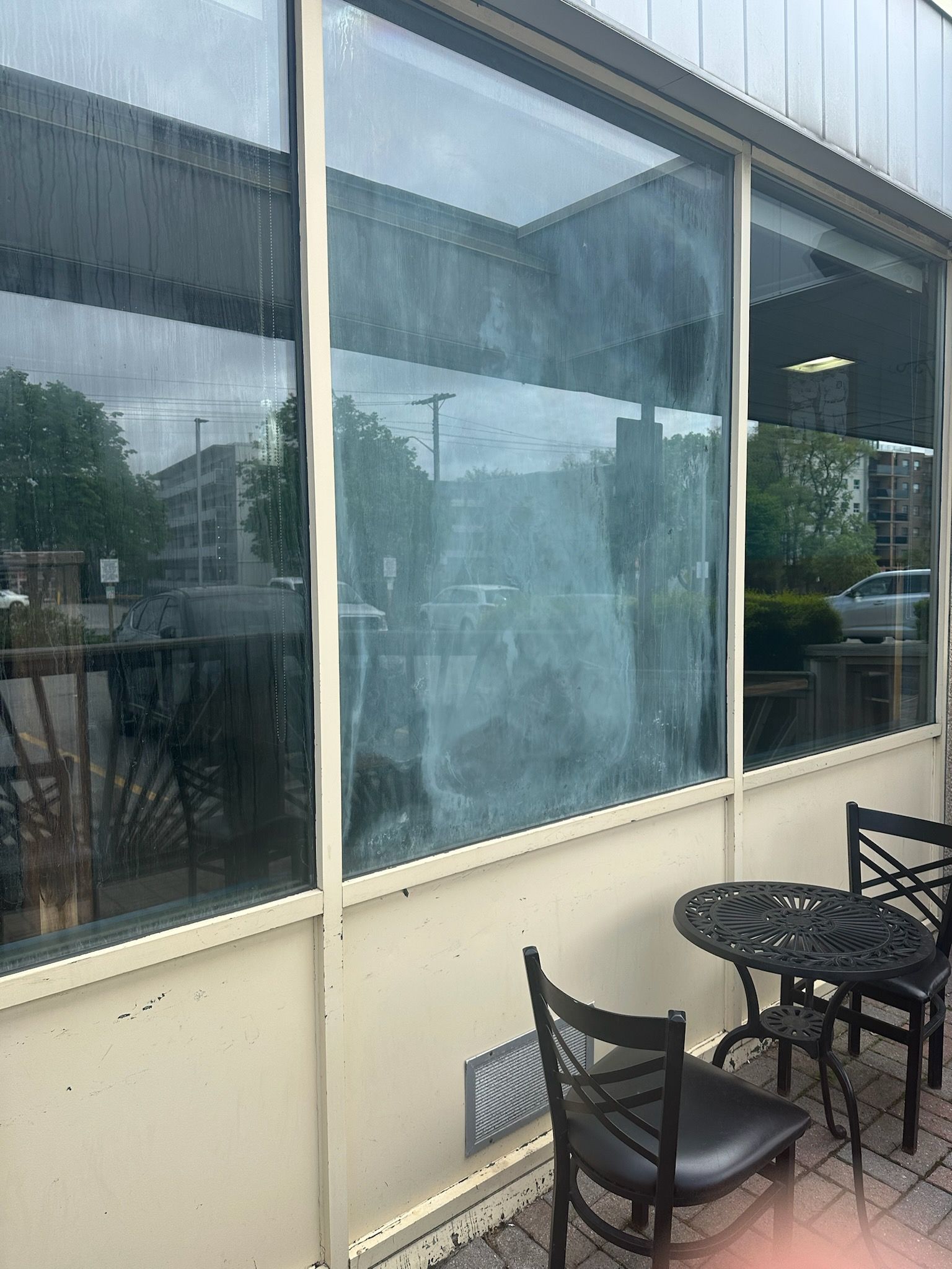 Outdoor café patio with black metal chairs beside large rain-speckled windows.