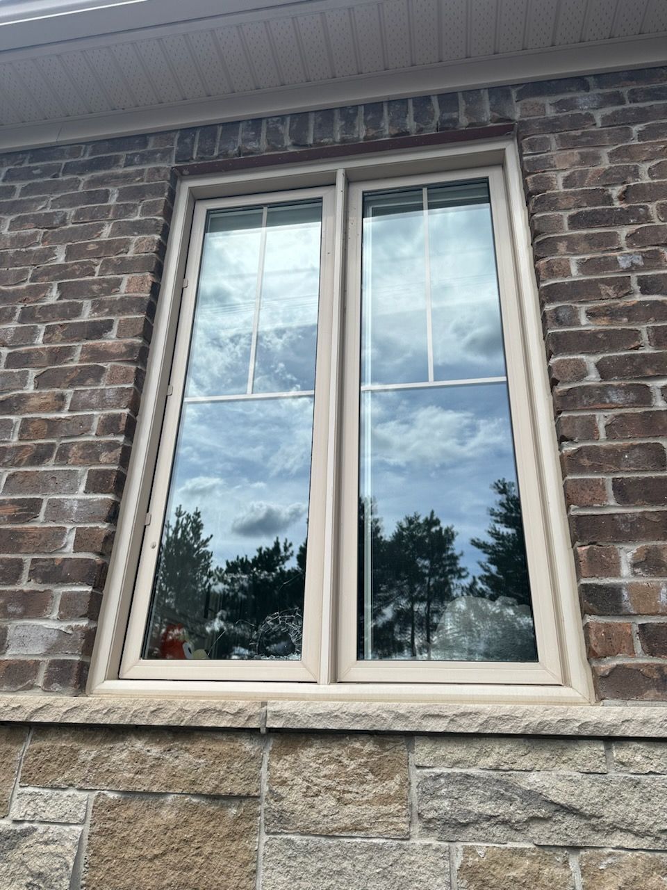 White-framed double window in a brick and stone house exterior, reflecting clouds and trees.