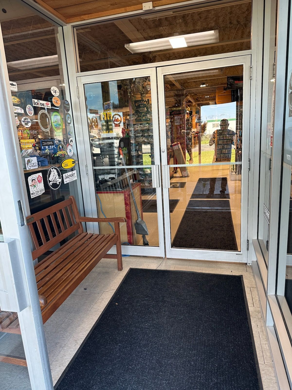 Storefront entrance with glass doors, a wooden bench, and a black doormat under a covered entryway.