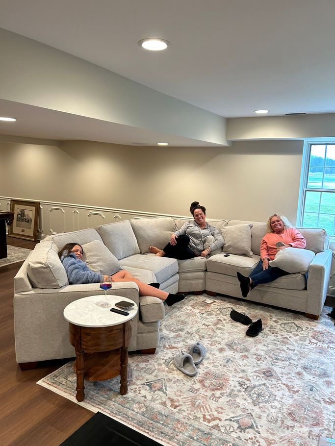Three people relaxing on a gray sectional in a bright basement with a patterned rug and coffee table.