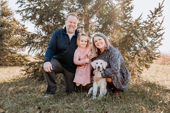 Family posing outdoors with a dog beneath tall trees on a sunny day