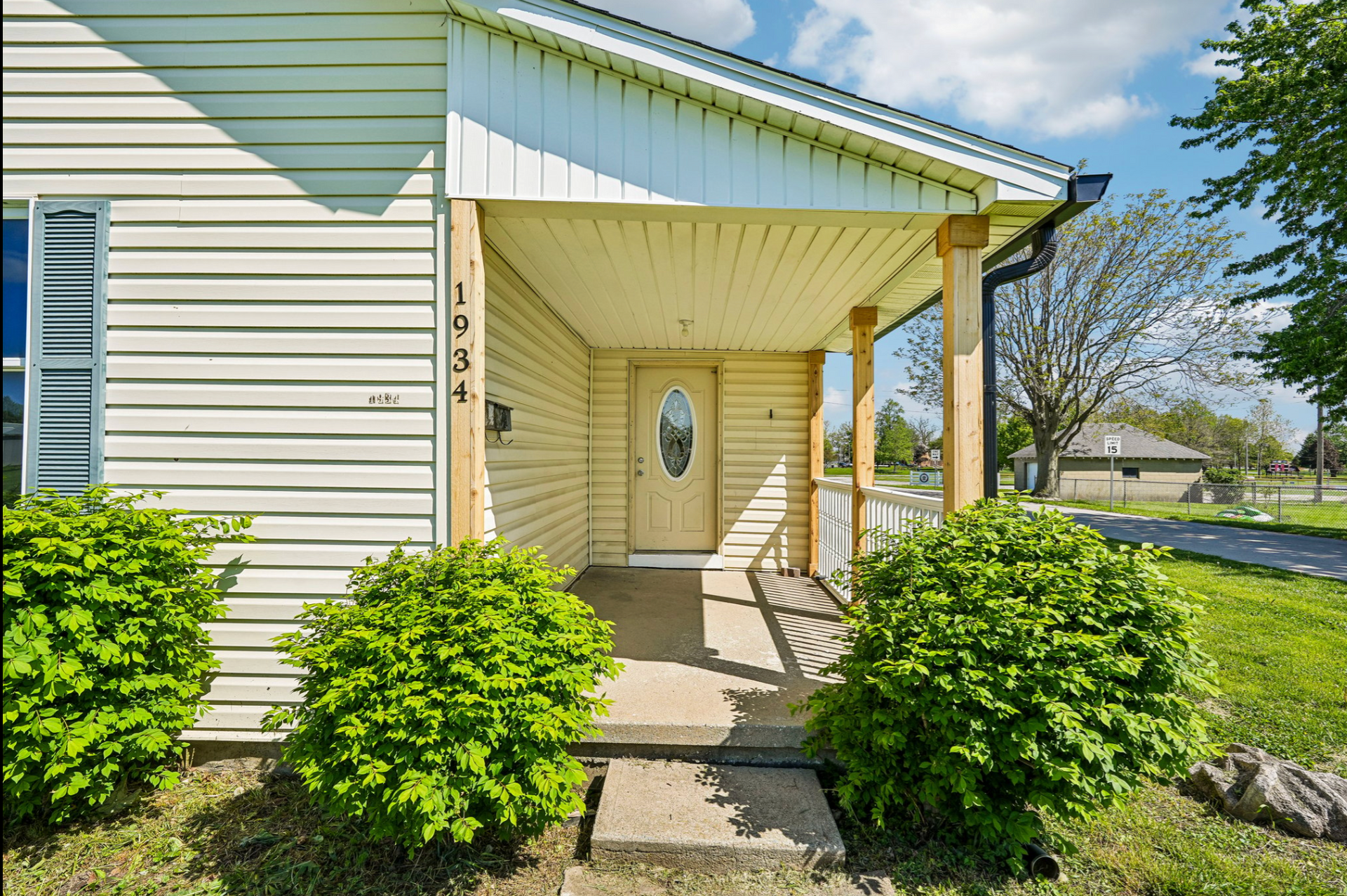 Front porch of a beige house with white trim, steps, and green shrubs in bright sunlight.
