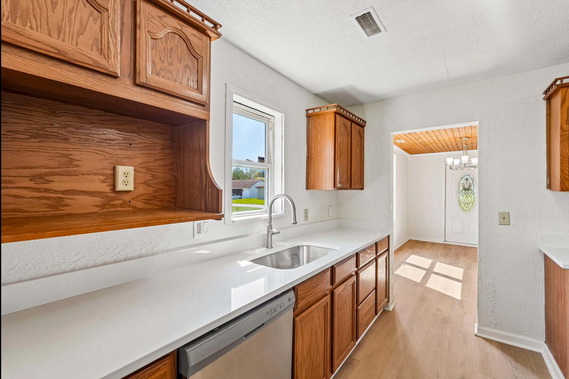 Bright kitchen with white counters, wood cabinets, sink by window, and sunlight on the floor.