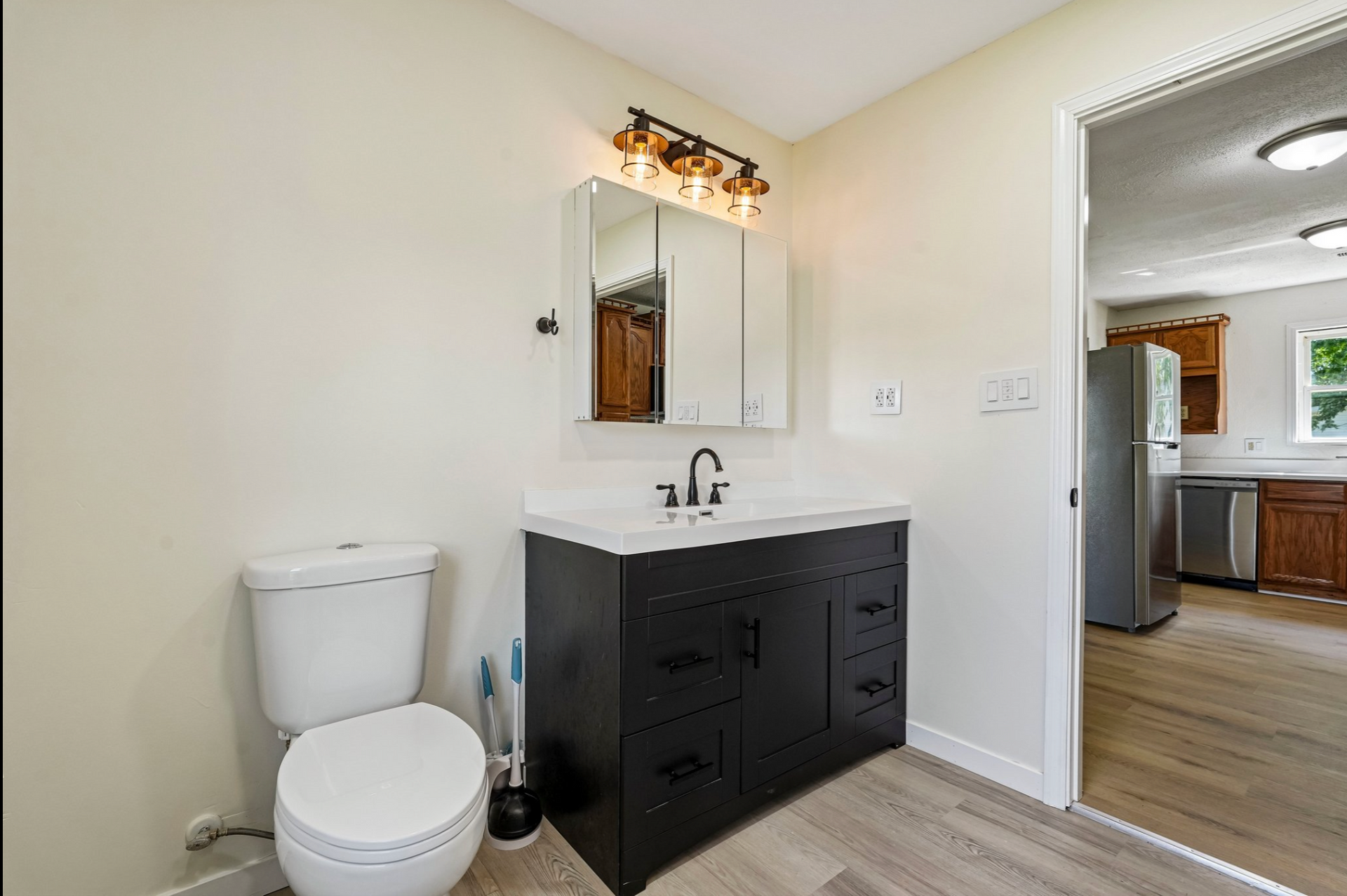 Small basement bathroom with white toilet and black vanity, mirror, and doorway to a kitchen.