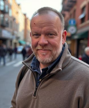 Smiling man in a gray fleece jacket standing on a blurred city street.