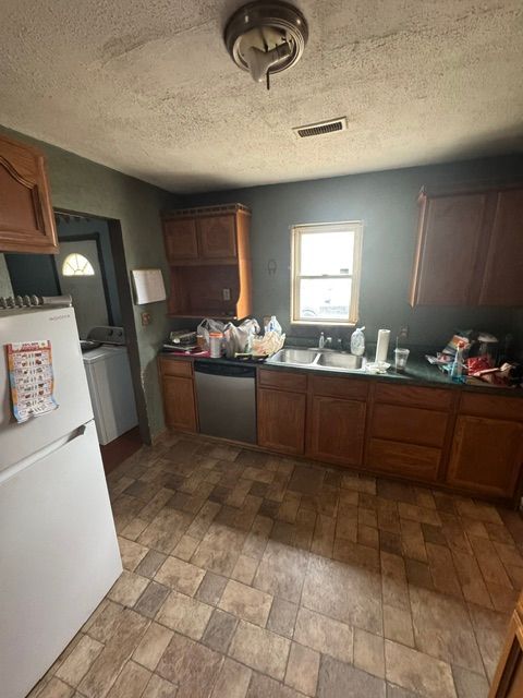Cluttered basement kitchen with brown cabinets, a white fridge, tiled floor, and dishes by a sunlit window