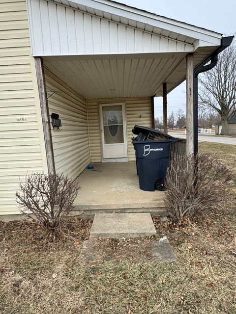Covered front porch with beige siding, a black trash bin, and shrubs beside a concrete walkway.