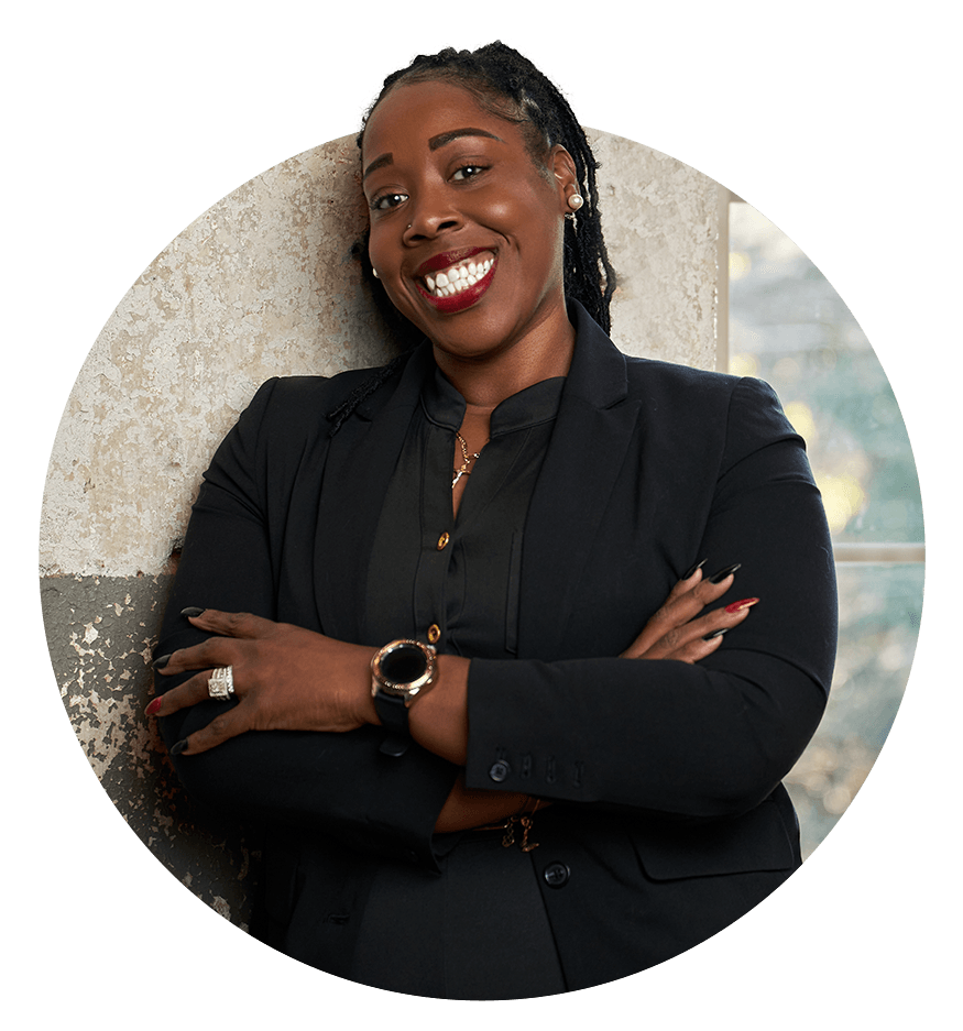 A woman in a suit is leaning against a wall with her arms crossed and smiling.