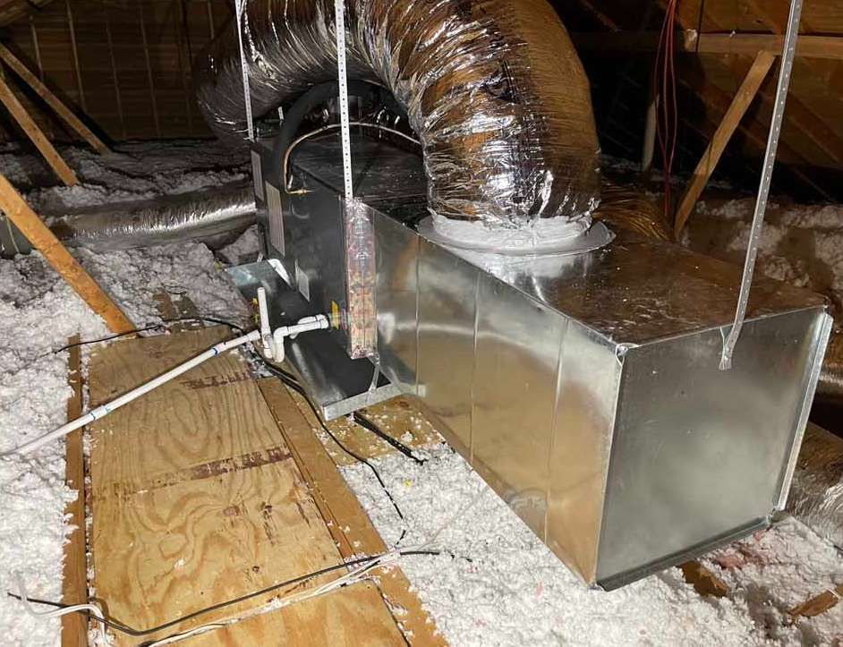 HVAC unit in an attic. Metallic ducts, insulation, wooden planks, and hanging supports are visible.