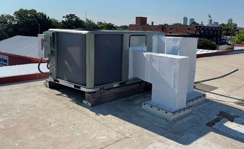 Rooftop HVAC unit with air ducts, on a flat rooftop, with city buildings in the background.