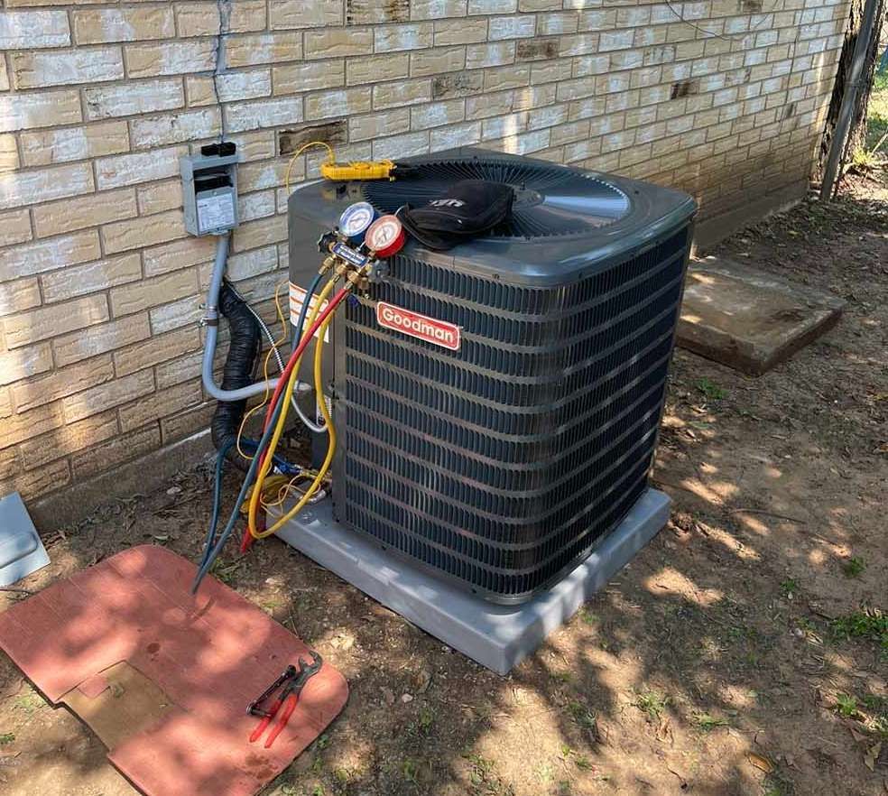 HVAC unit being serviced outdoors. Gauges are connected, tools nearby, brick wall background.
