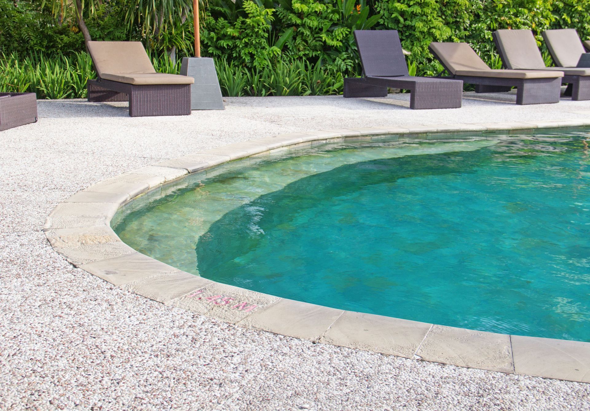 Curved swimming pool with turquoise water beside a white gravel deck and lounge chairs.