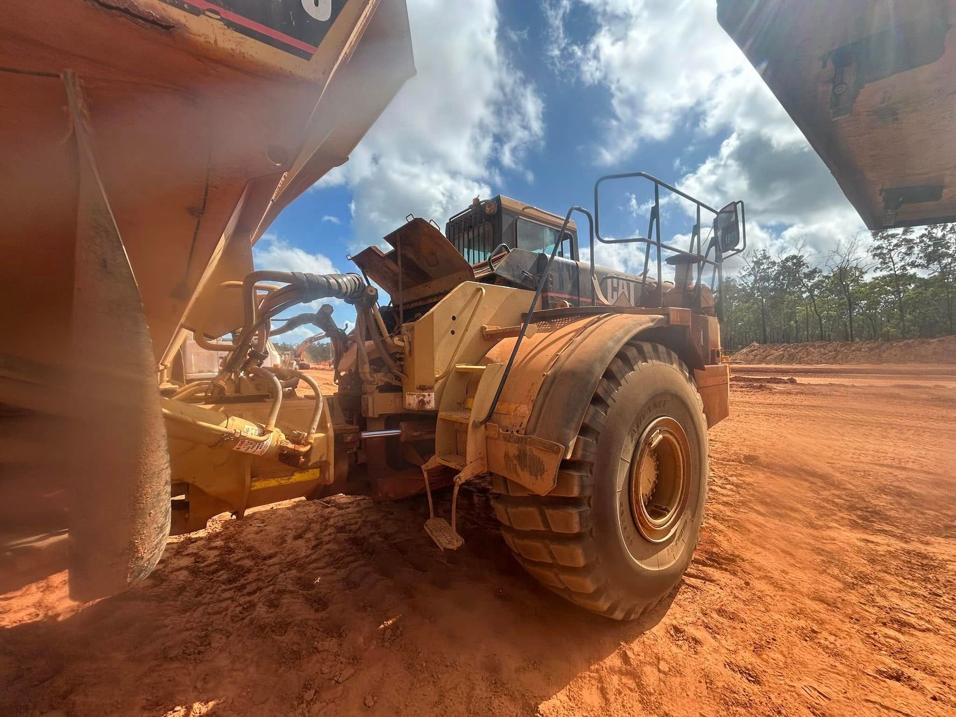 Yellow earthmover vehicle on red dirt under a blue sky, partially obscured by a larger vehicle — Full Noise Mechanical in Humpty Doo, NT