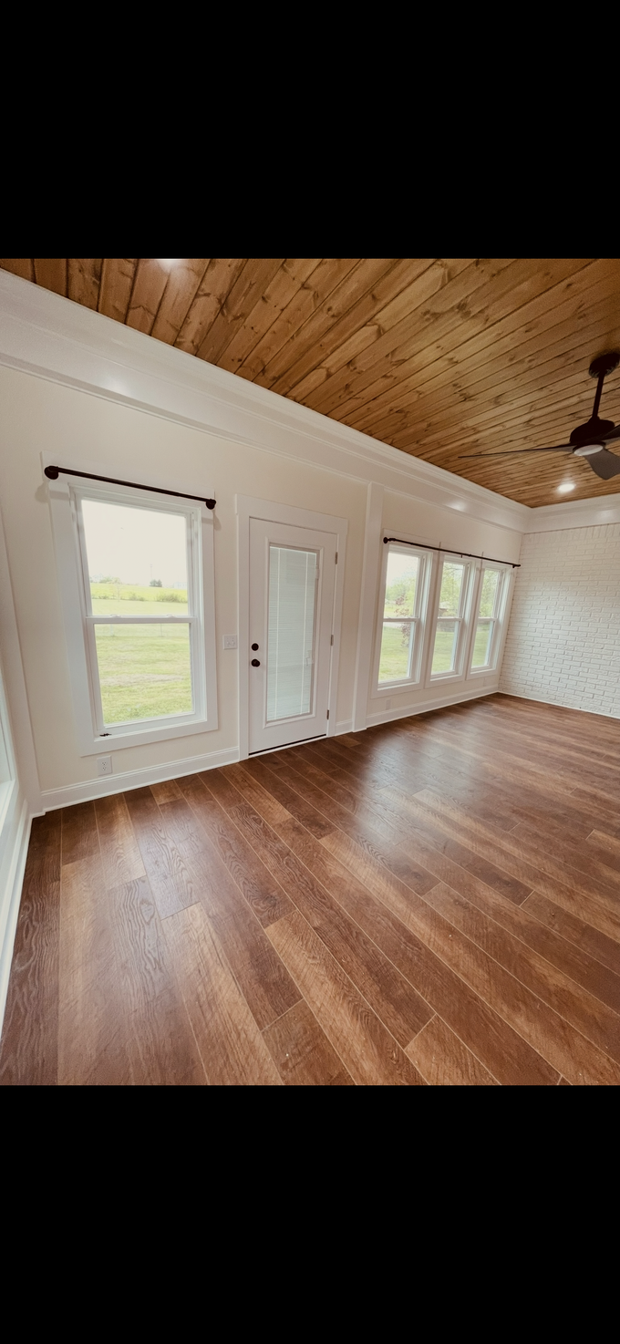 Empty room with wood floors, white walls, ceiling fan, and large windows showing greenery outside